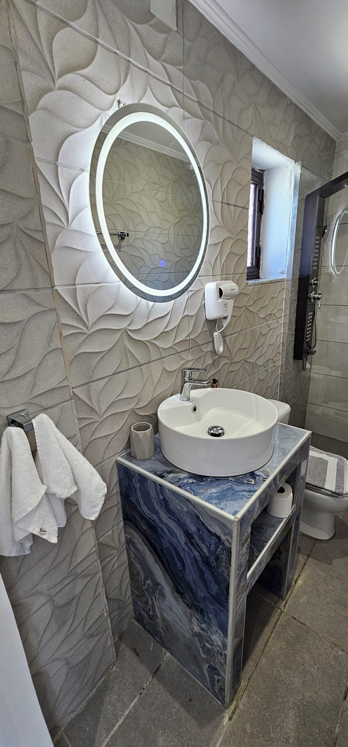Un baño con tocador de mármol, un espejo redondo, toallero y ducha de mano.