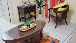 A living room with a glass-top coffee table, a wicker chair, a potted plant, a television, and a colorful rug.