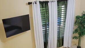 A hotel room with a bed, a TV mounted on the wall, a window with curtains, and a potted plant.
