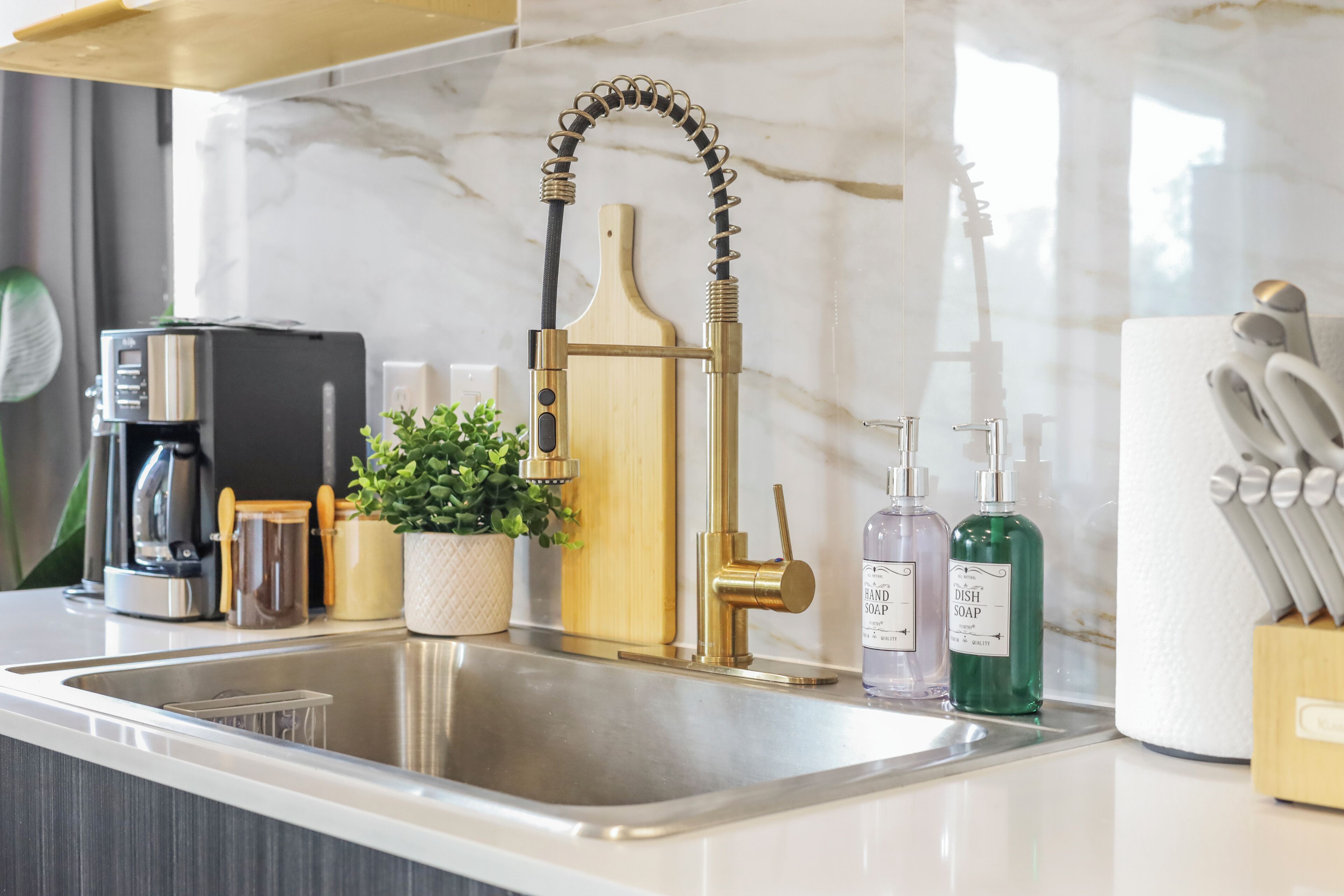 A modern kitchen with a stainless steel sink, a gold-colored faucet, a coffee machine, and various kitchen utensils.