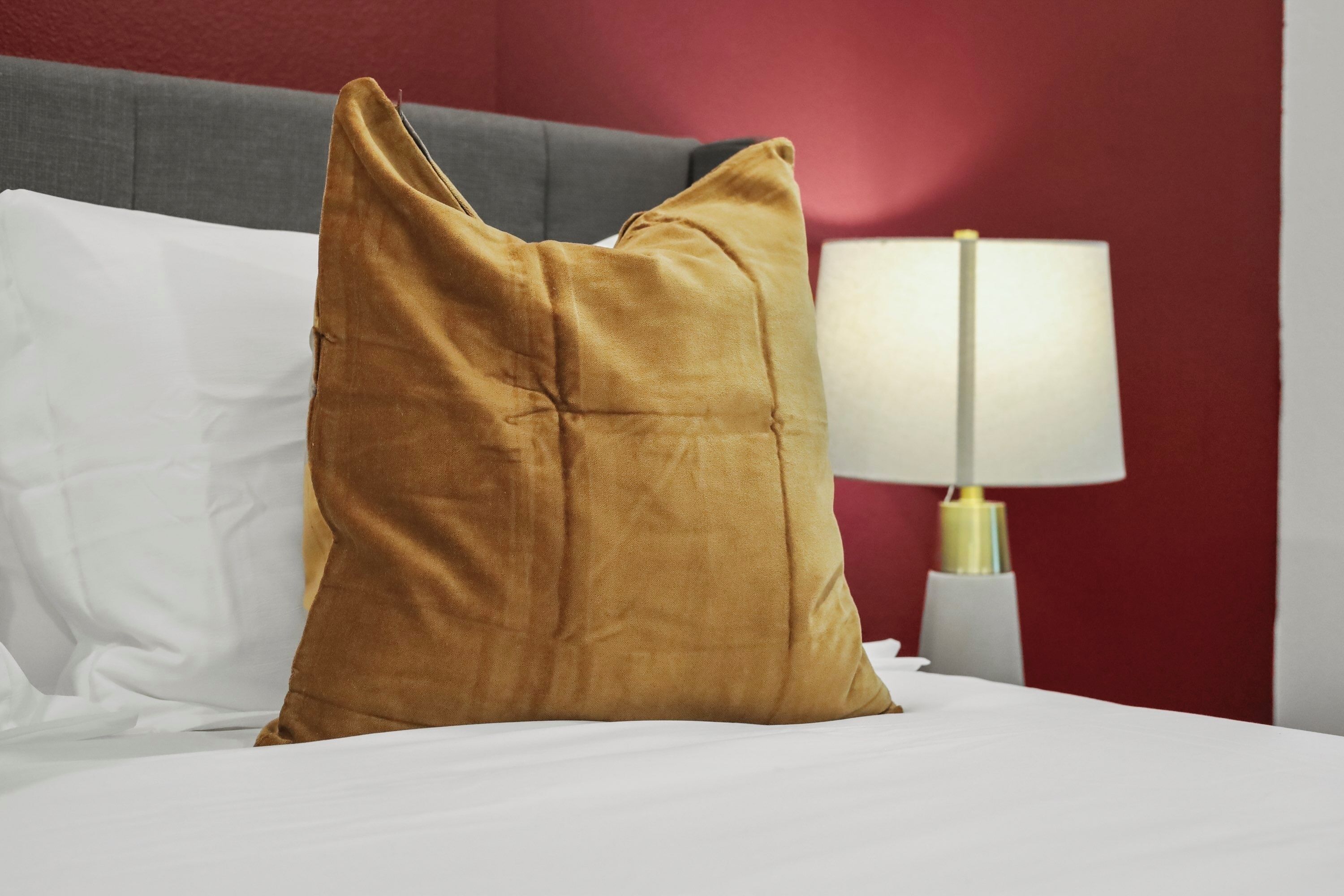 A bed with white linens, a mustard-colored pillow, a gray headboard, and a bedside lamp with a white shade and gold accents.