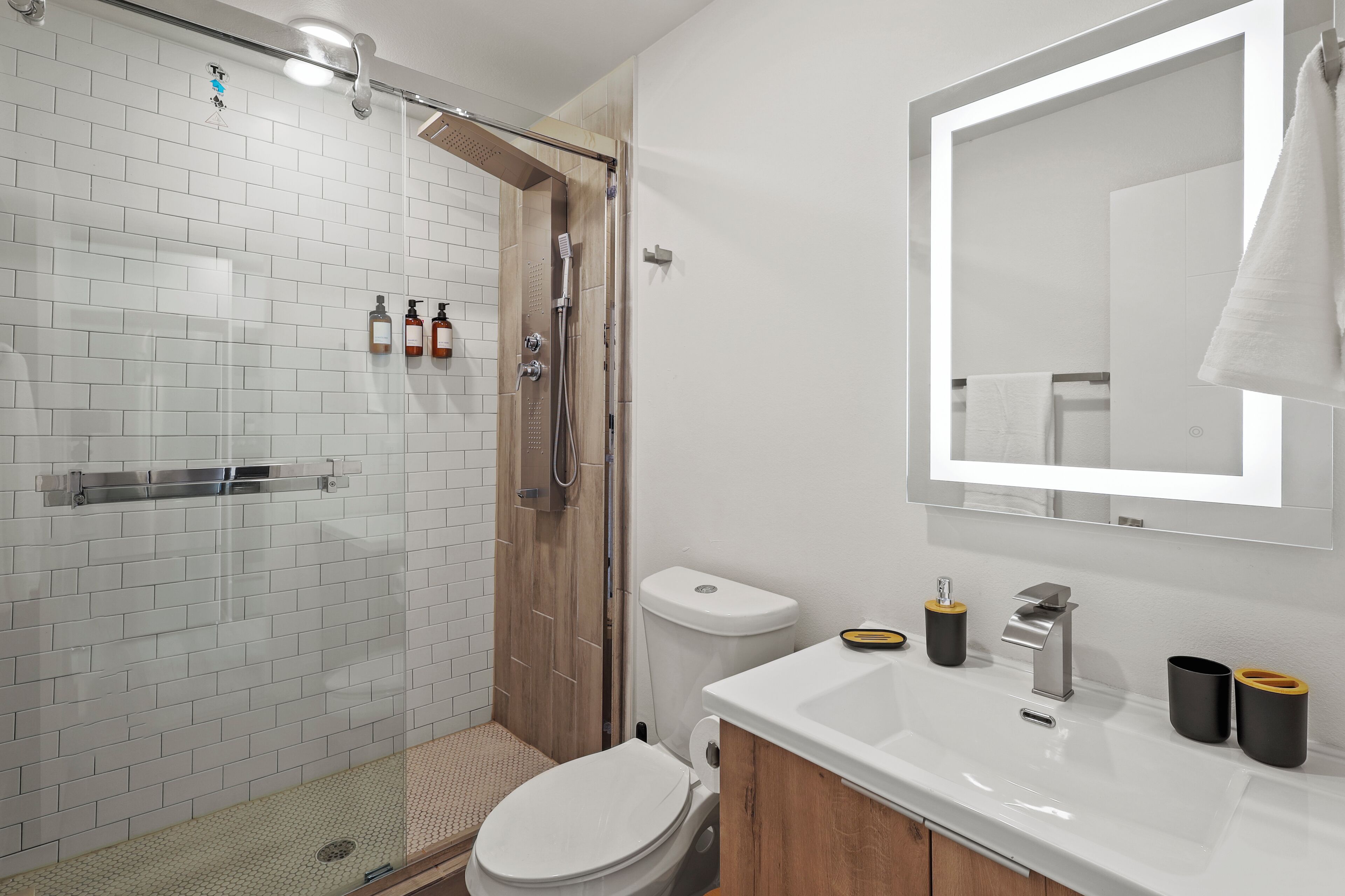 Ein modernes Badezimmer mit Duschkabine aus Glas, weißen Fliesen, einer weißen Toilette und einem Waschbecken mit schwarzen und goldenen Accessoires.