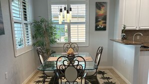 A dining area with a glass table and metal chairs, a potted plant, and a window with blinds.