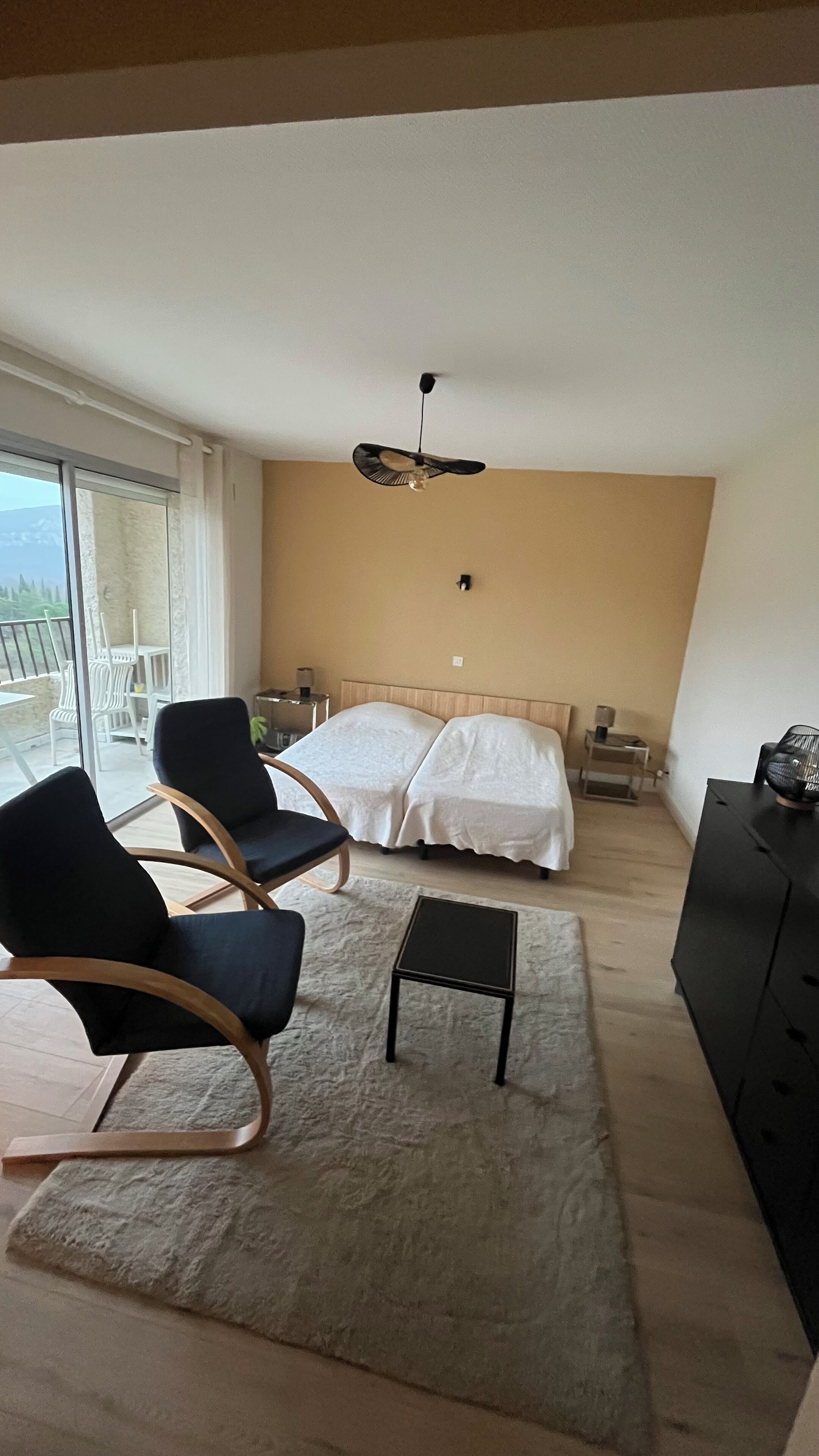 A bedroom with a bed, two chairs, a small table, a ceiling fan, and a view through a large window.