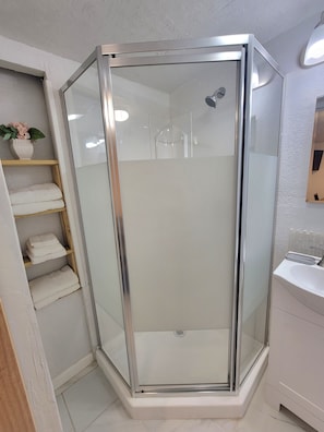 Eine Eckdusche mit Glastür, ein Holzregal mit Handtüchern und ein weißes Waschbecken.