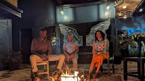 Three people seated around a campfire in an outdoor setting with stone flooring and a rustic interior. - Idembe Heritage (Nyamabuye)
