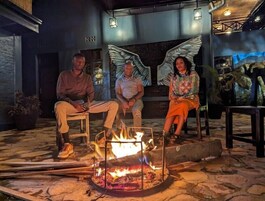 Tres personas sentadas alrededor de una fogata en un entorno al aire libre, con suelo de piedra y un interior rústico.