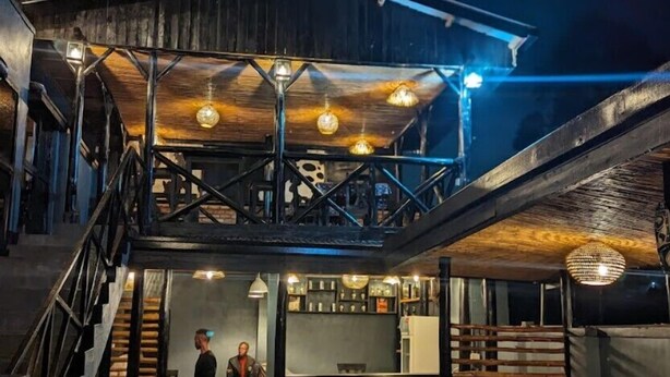 A rustic interior with a wooden staircase leading to a mezzanine level, a bar area with shelves, and hanging pendant lights. - Idembe Heritage (Nyamabuye)