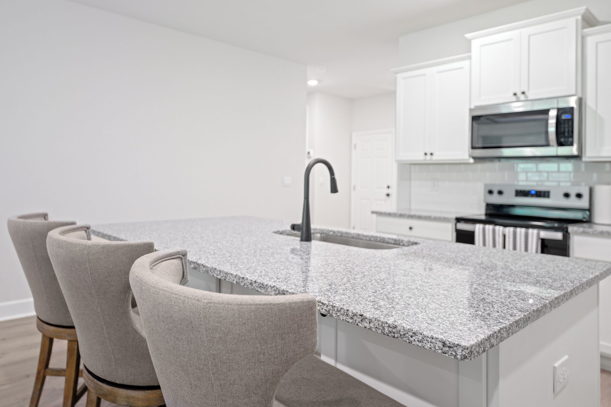 A modern kitchen with white cabinets, a granite countertop, a stainless steel microwave, and a built-in oven. There are three upholstered chairs with wooden legs.
