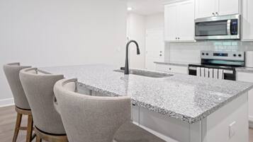 A modern kitchen with white cabinets, a granite countertop, a stainless steel microwave, and a built-in oven. There are three upholstered chairs with wooden legs.