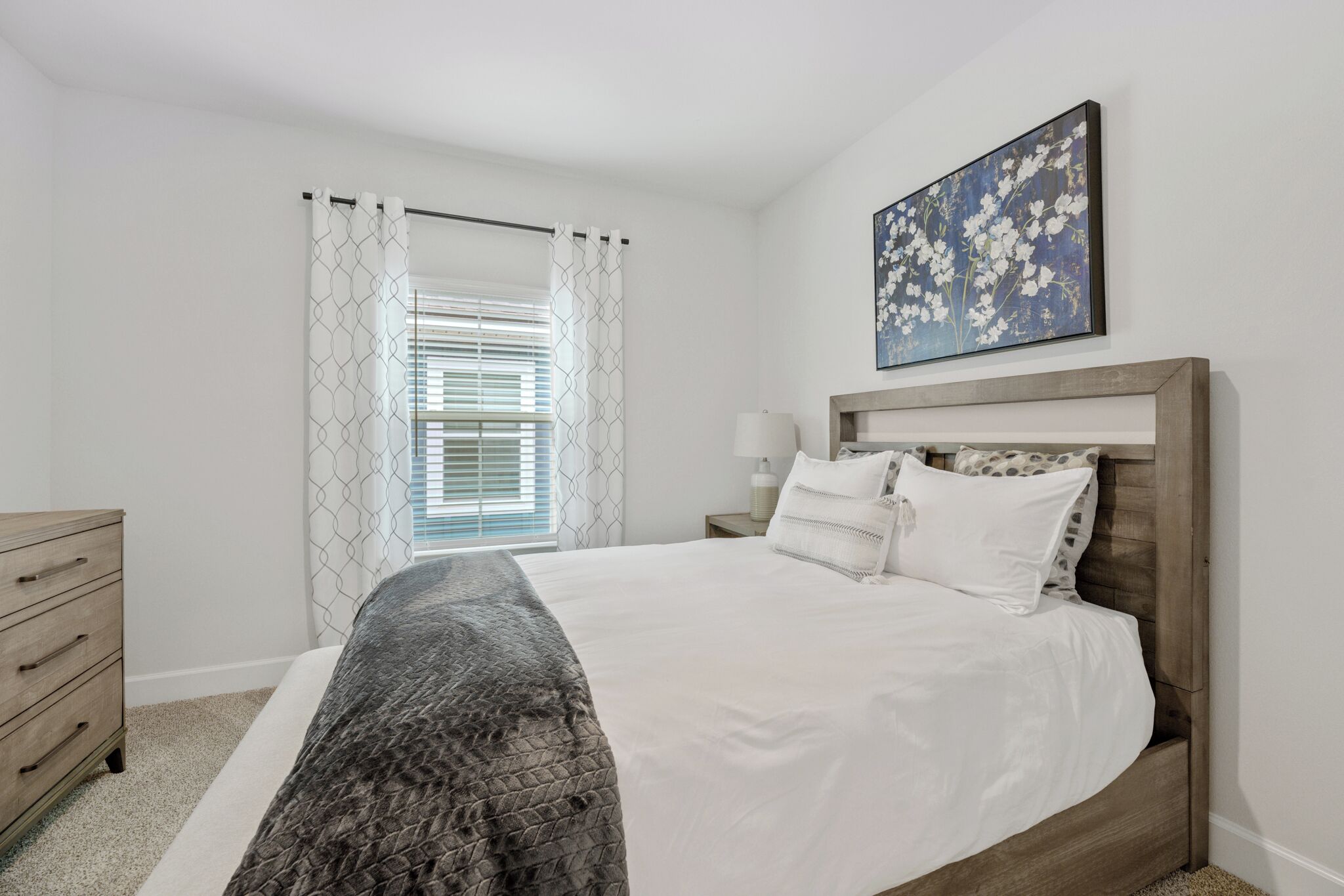 A neatly arranged bedroom with a bed, a dresser, a bedside table with a lamp, and a window with blinds.