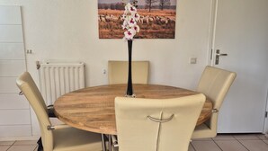 Et rundt træbord med fire beige stole, en vase med blomster og et maleri af en mark med får på væggen.