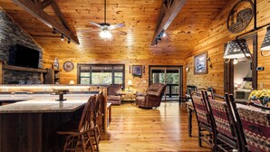 A wooden cabin interior with a stone fireplace, a bar area, and a dining space with patterned chairs. - Trails End Lodge - 5 BR cabin in Morganton with Games, WiFi, Hot Tub (Morganton)