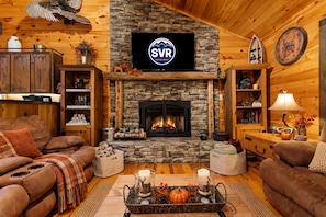 L’intérieur chaleureux d’un chalet avec une cheminée en pierre, des murs en bois, un téléviseur fixé au mur et une bibliothèque.