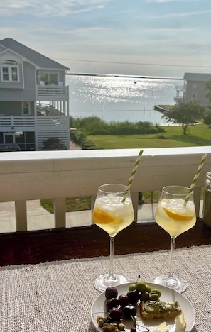 A balcony with two glasses of a light-colored beverage, a plate of olives and a slice of pie, and a view of a house and water.