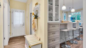 Un pasillo con un banco, una puerta blanca y una zona de cocina con taburetes de bar y luces colgantes.