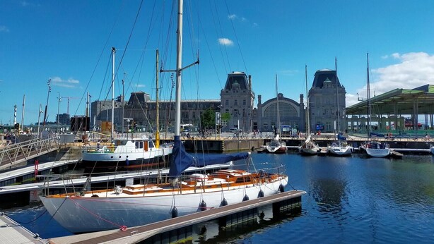 Una marina con varios barcos amarrados, un edificio grande de arquitectura distintiva al fondo y un cielo azul despejado.