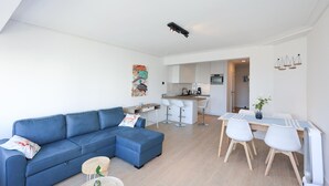 Una sala de estar moderna con un sofá seccional azul, una mesita redonda con un florero y una cocina con armarios blancos y un taburete de bar.