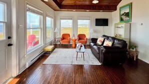 A living room with a wooden ceiling, two windows with blinds, a black leather sofa, an orange armchair, a white rug, and a television on the wall. - Escape to your private oasis in our ocean front home with breathtaking views.  (Grand Bank)