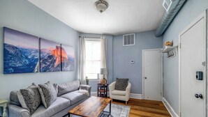 A modern living room with a grey sofa, a wooden coffee table, and a white armchair. There are two large paintings on the wall, a window with curtains, and a door with a coat rack. - Perfect Adams Morgan Location! Charming & Historic (Washington)