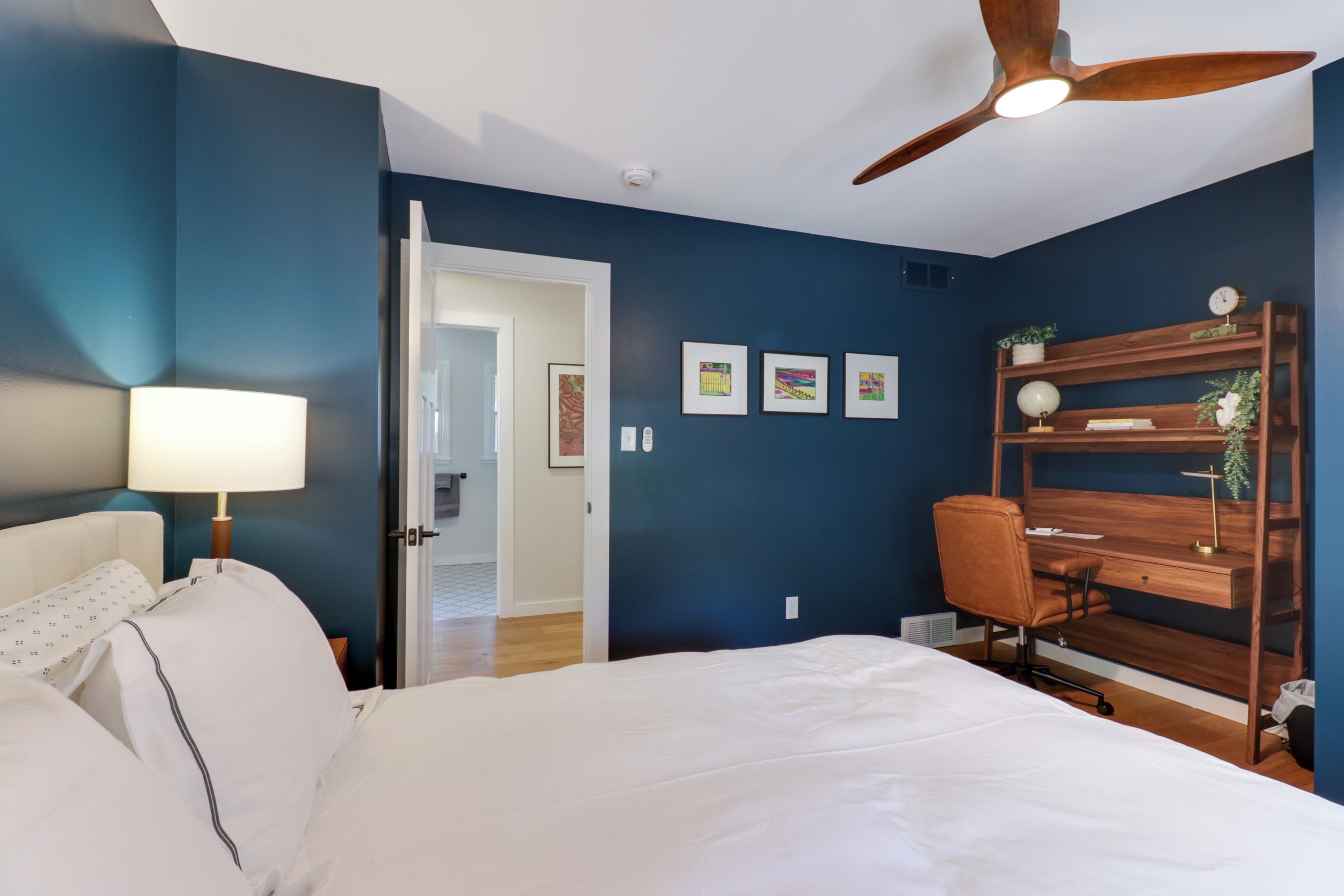 A bedroom with a bed, a desk, a chair, a ceiling fan, and artwork on the wall.