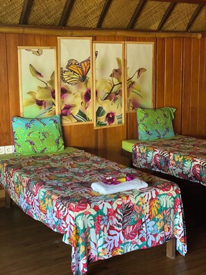 Ein Zimmer mit zwei Betten, jedes mit einem blumigen Überwurf, einem grünen Kopfkissen und einem hölzernen Kopfteil. An der Wand hängt ein Blumenbild und ein Schmetterling.
