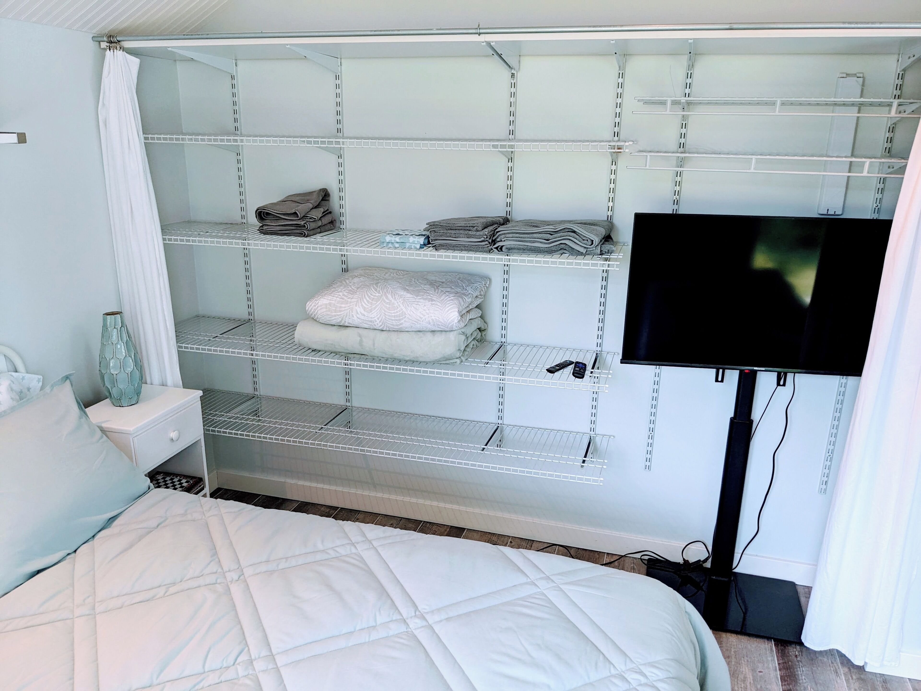 A bedroom with a bed, a TV mounted on the wall, and a built-in shelving unit with folded towels and blankets.