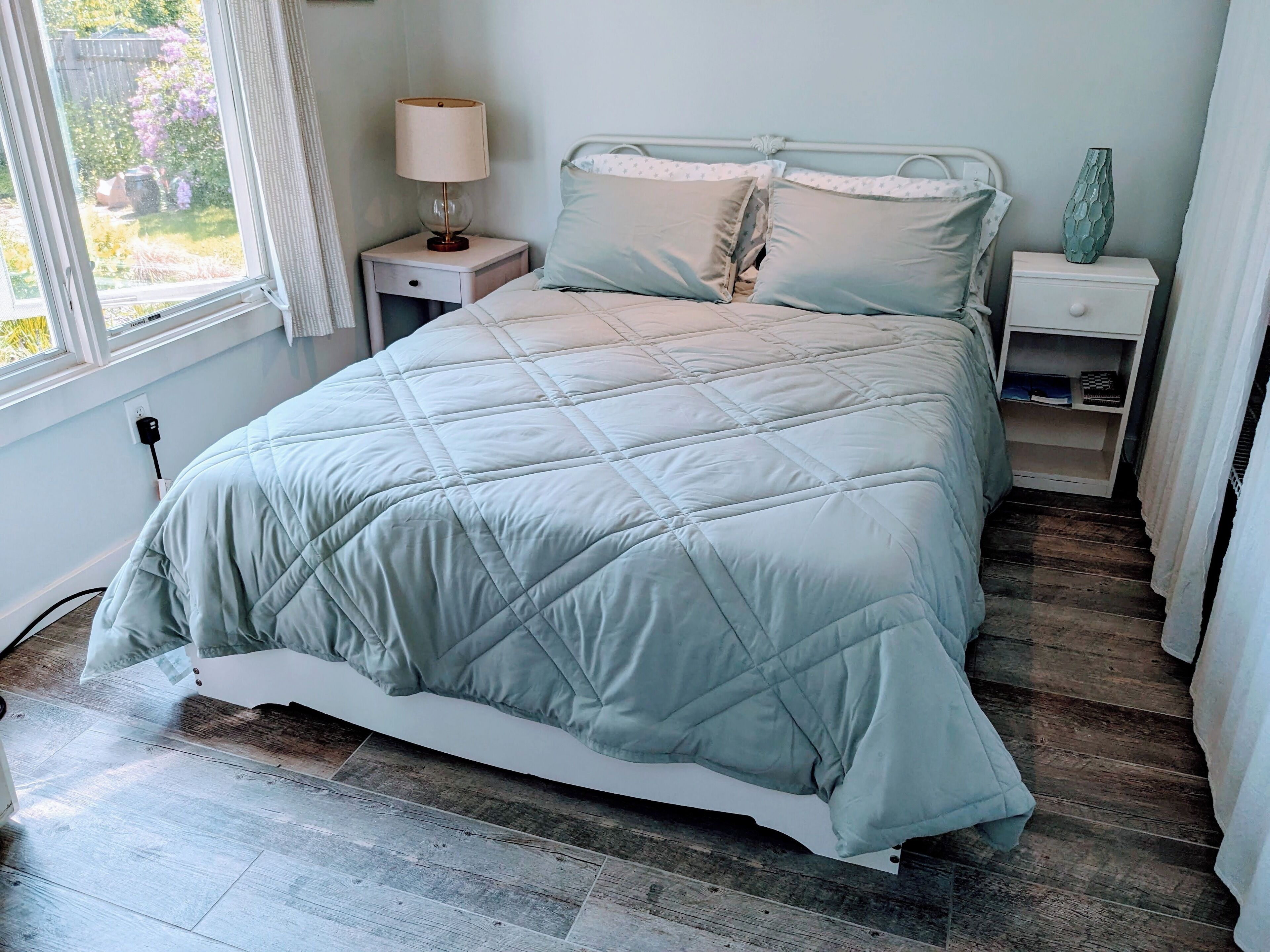A bedroom with a bed, a nightstand, a lamp, a vase, and a view of the outdoors.
