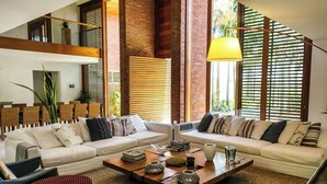 Ein geräumiges Wohnzimmer mit einer großen Sofagarnitur, einem Couchtisch mit Dekoration und großen Fenstern mit Jalousien.