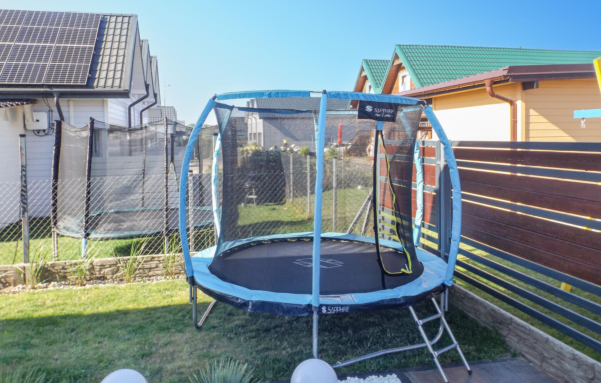 Un trampoline doté d’un filet de sécurité, installé dans un jardin avec un mur en pierre et une clôture en bois.
