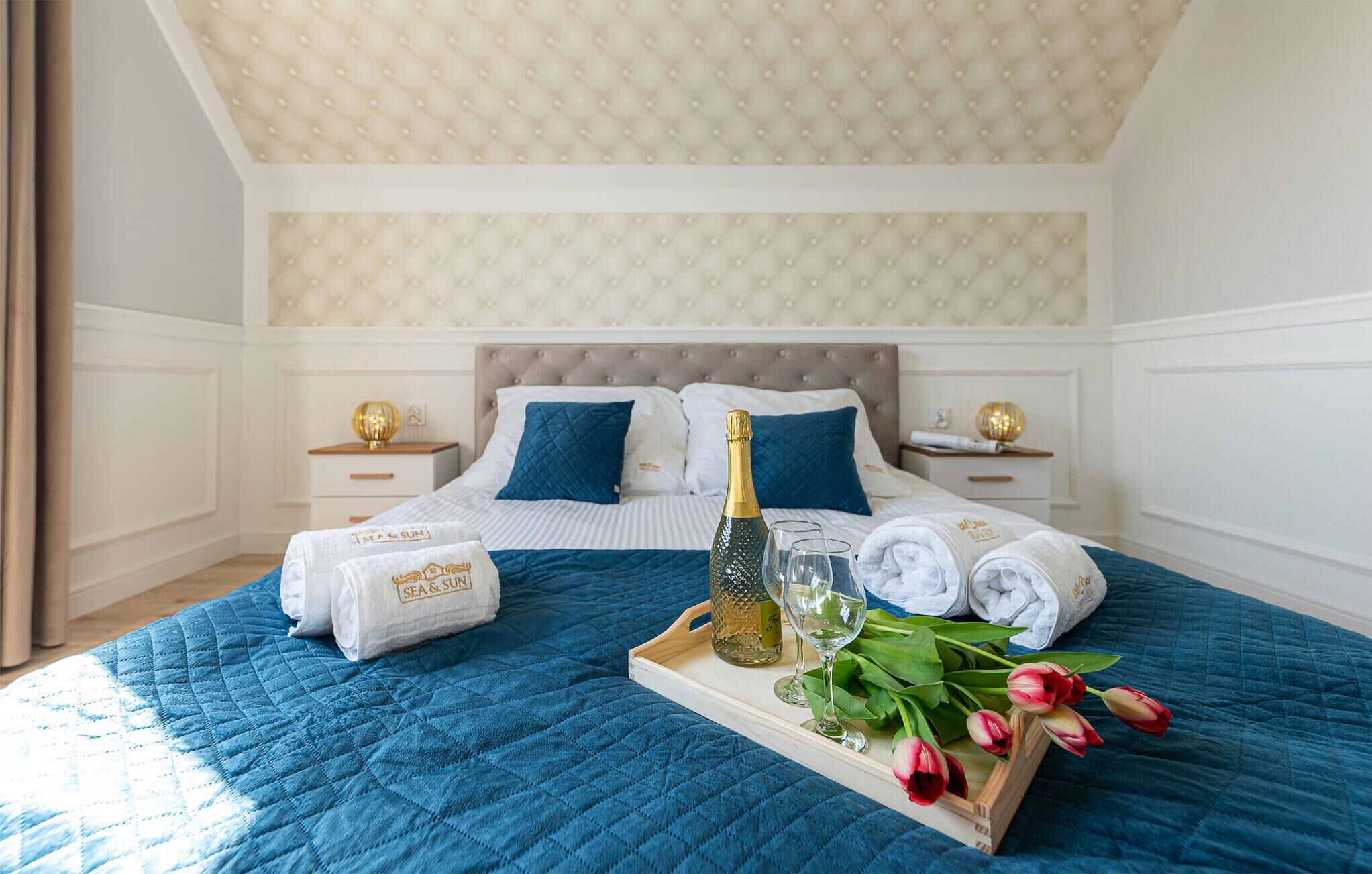 Un lit bien fait, recouvert d’une couette bleue, avec deux oreillers, une bouteille de champagne et un plateau contenant des serviettes roulées et des tulipes.