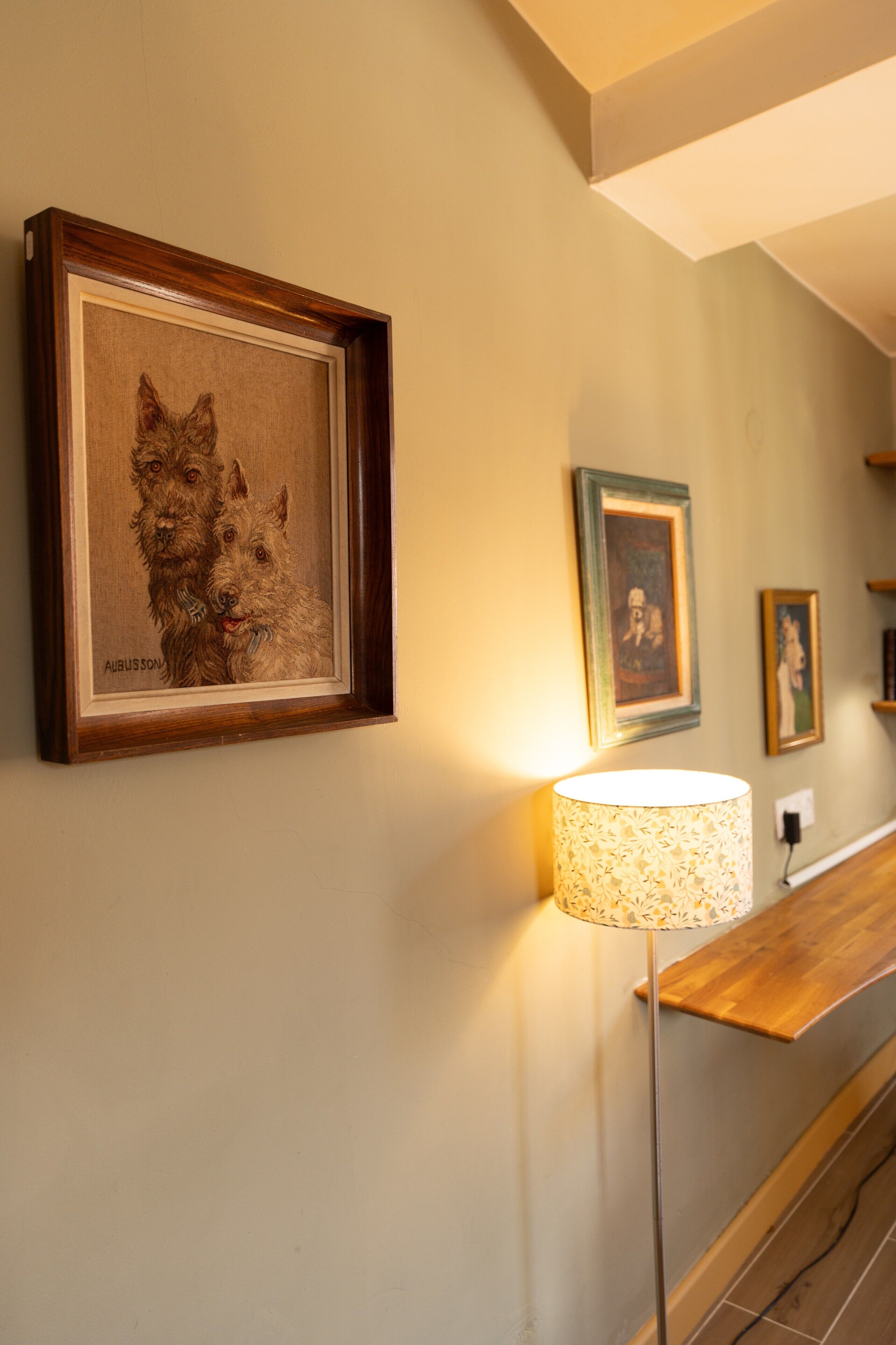 Una obra de arte enmarco muestra dos perros en una pared, una lámpara de pie y un banco de madera.