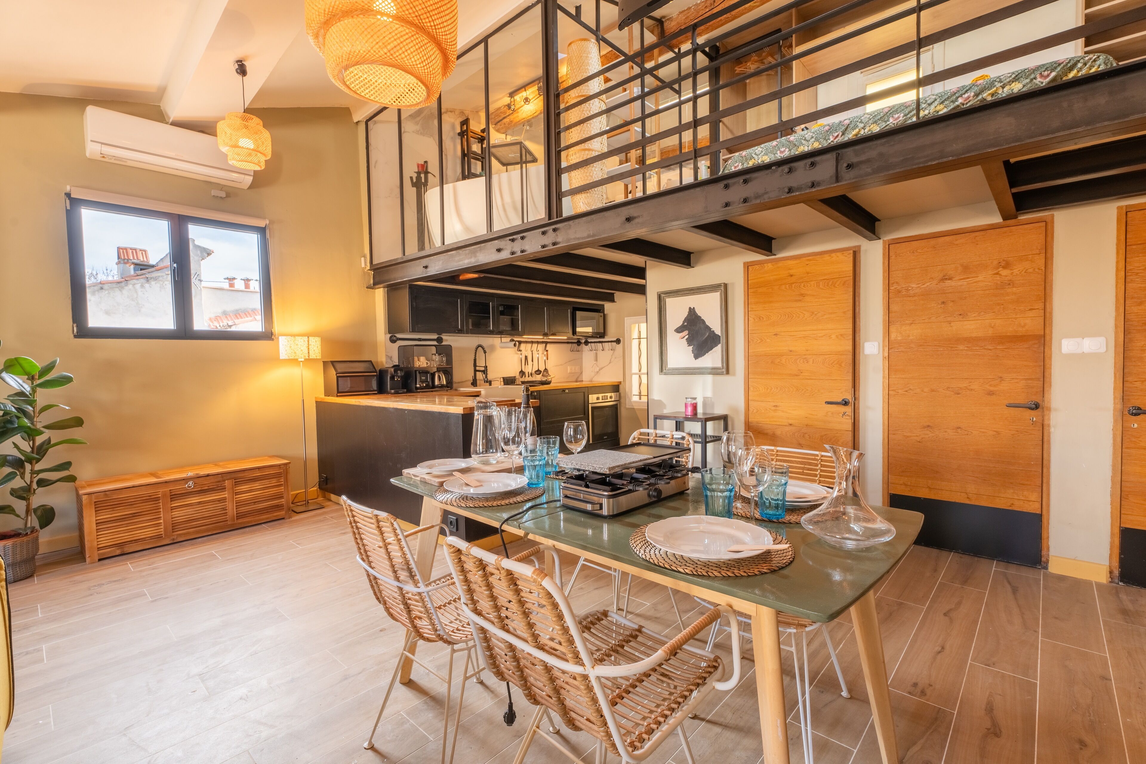 A modern kitchen with a dining area, featuring a high ceiling, a staircase, and a large window.