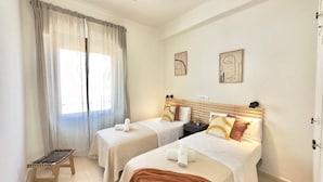 Habitación de hotel con dos camas, ventilador de techo, una banca y obras de arte en las paredes.