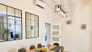 Un comedor con una mesa larga, sillas y un bol de limones. Hay dos espejos montados en la pared, una lámpara en el techo y una puerta con paneles de vidrio esmerilado.