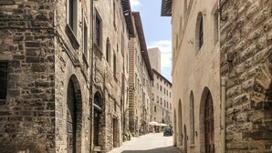 A narrow street lined with historic stone buildings, featuring arched doorways and windows, leading towards a distant open area with people and a parked car. - Lovely apartment in Gubbio with WiFi (Gubbio)