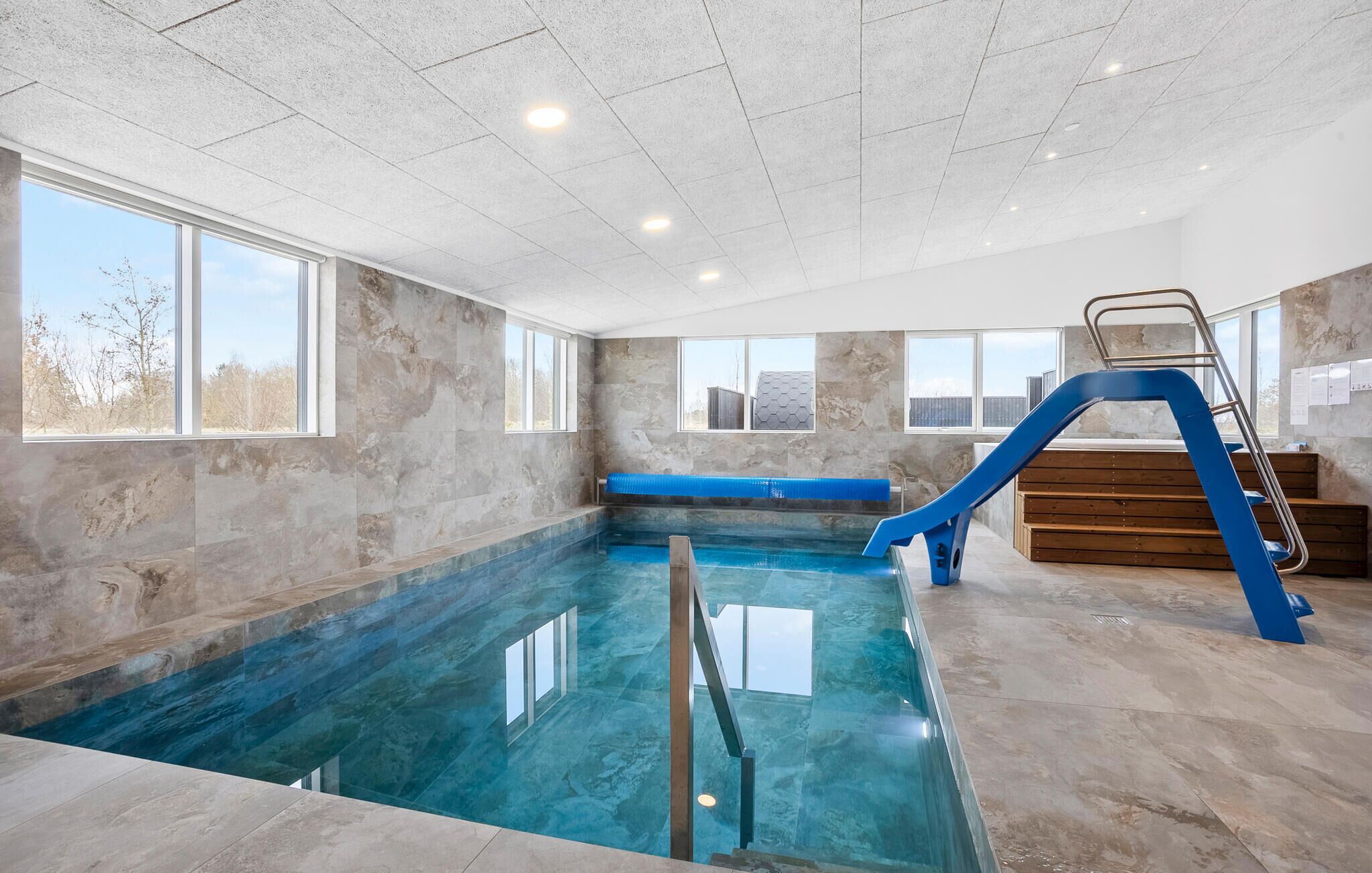An indoor swimming pool with a blue slide, stone walls, and large windows.