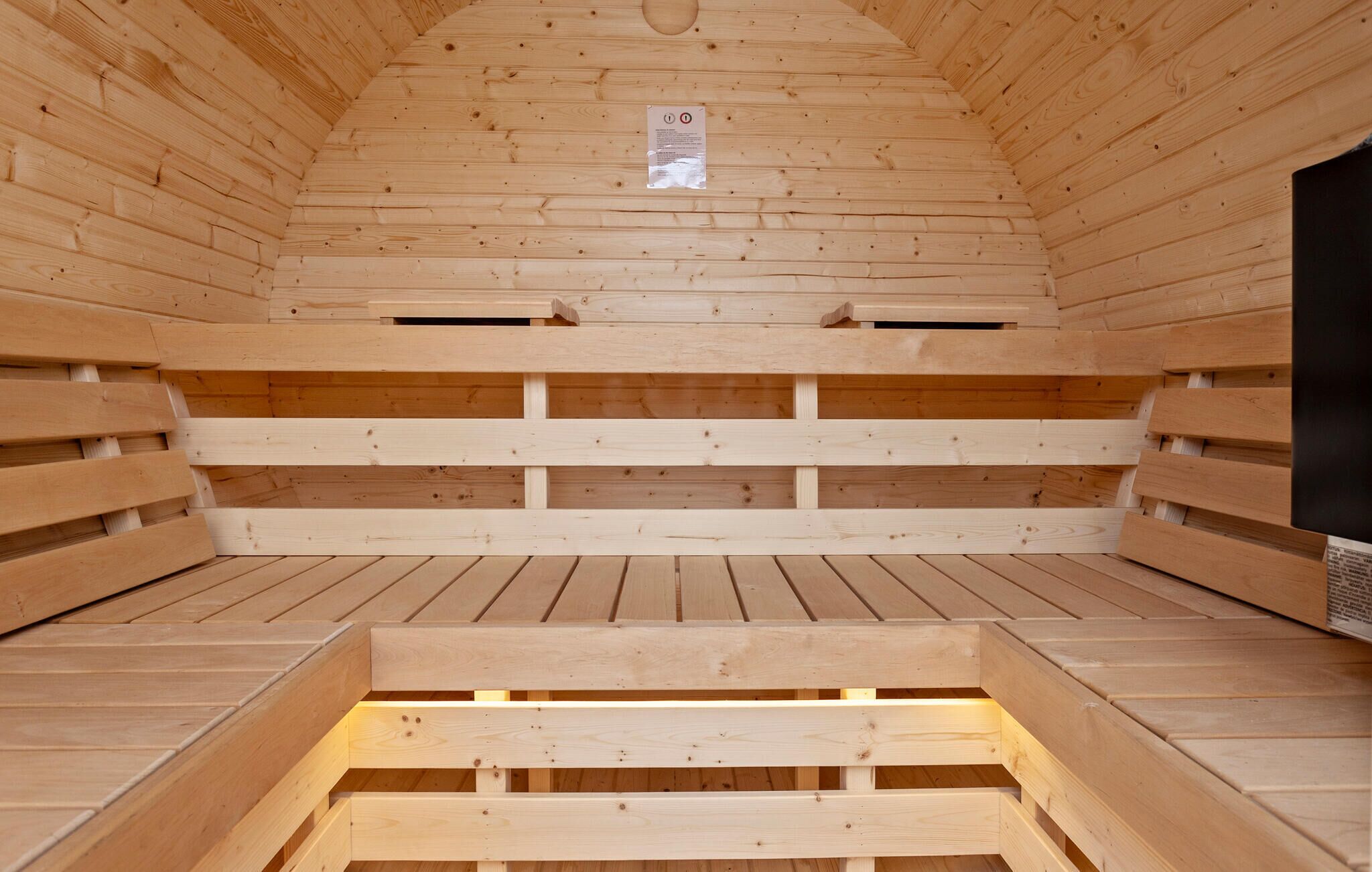 Un sauna avec des bancs en bois disposés en arc de cercle, un petit trou d’aération en haut et un objet noir sur le côté droit.