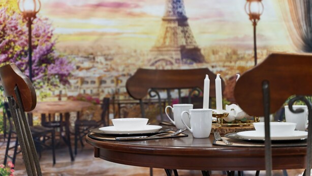 A dining table set with white plates, cups, and a candle, with a backdrop of the Eiffel Tower and a cityscape. - Pixar Pier: Heated Pool, Arcade, Golf, and More!+ (Garden Grove)