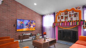 A living room with a brick wall, a flat-screen TV, orange sofas, a wooden coffee table, and a fireplace decorated with orange flowers. - Pixar Pier: Heated Pool, Arcade, Golf, and More!+ (Garden Grove)