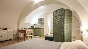 A bedroom with a large bed, a small table, and a chair. There is a kitchenette in the background with cabinets and a sink. - Tea Tree Apartments 1 (Castellana Grotte)