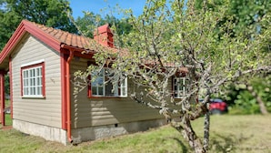 Ein kleines Holzhaus mit rotem Dach und weißen Fensterrahmen, umgeben von Gras und Bäumen.