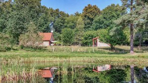 Ländliche Landschaft mit einem Teich, der ein Gebäude mit rotem Dach spiegelt, umgeben von Bäumen und Vegetation.