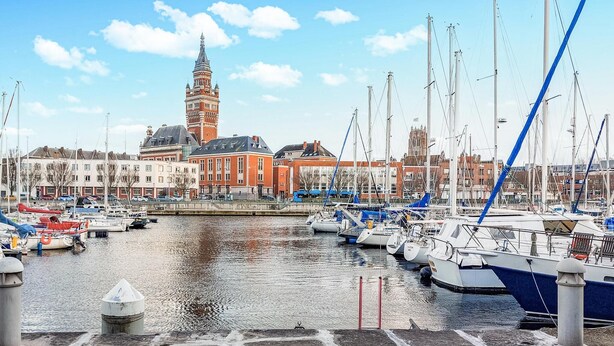 Marina com barcos atracados, uma torre imponente e edifícios ao fundo.