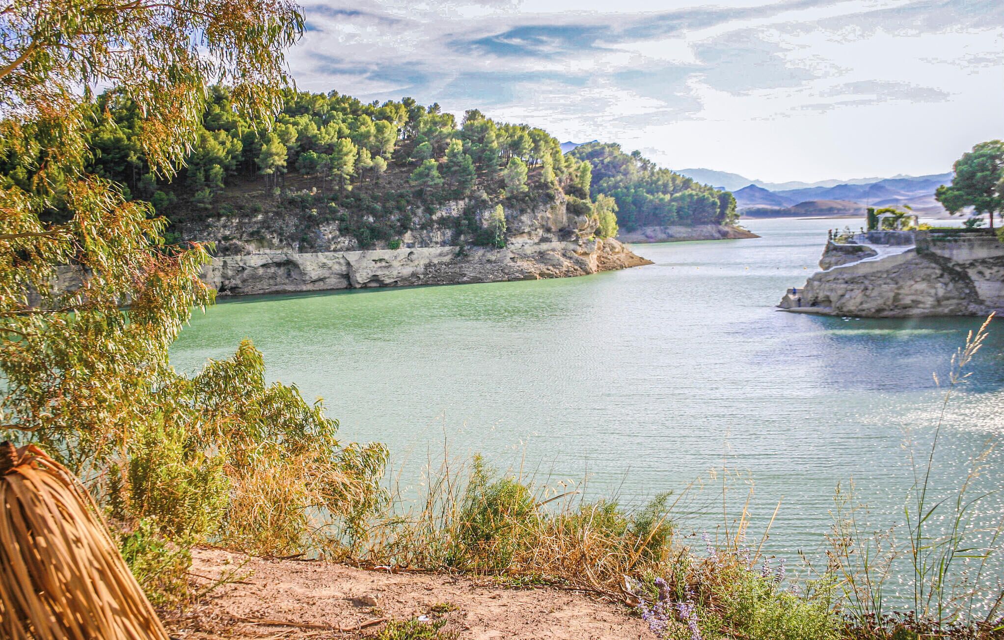 A coastal landscape with a calm lake, rocky cliffs, and dense greenery.