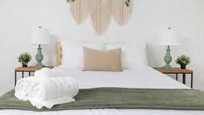 Een netjes opgemaakt bed met witte lakens, een groene plaid en een macramé wanddecoratie erboven.