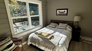 Une chambre à coucher avec un lit, un ventilateur de plafond, une fenêtre donnant sur des arbres, un fauteuil en osier et une table de chevet avec une lampe.