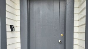 A grey door with a brass doorknob and a keyhole, set in a white-framed entrance with a small intercom on the left. - Magpie Cottage. Newly Renovated , 2 Bed, 2 bathroom close to Napier CBD (Napier)