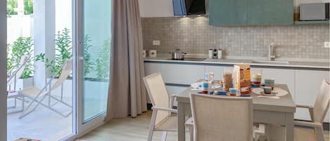 A modern kitchen with a dining area, featuring a large window, sliding doors, and a view of greenery.