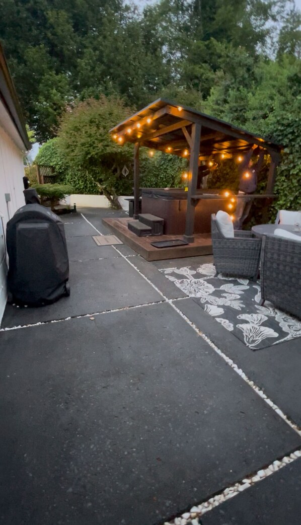 Un patio cubierto con mobiliario exterior, una pérgola con luces colgantes y un pequeño jardín.